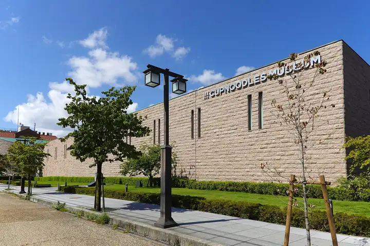 Stone exterior of the CUPNOODLES MUSEUM building with a hedge, trees and a lamppost along the pedestrian path