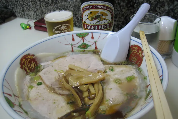 A bowl of Onomichi ramen with sliced pork, bamboo shoots, and scallions in soy sauce broth, served with beer