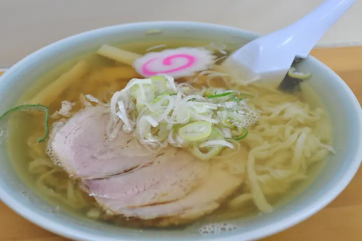 A bowl of clear soy sauce ramen with curly noodles, sliced pork, narutomaki, and chopped scallions in a blue bowl