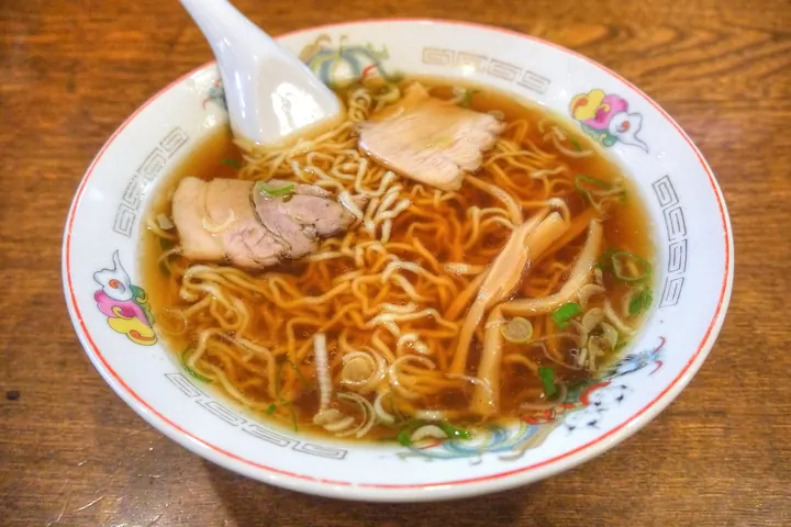 A bowl of soy sauce ramen with thin curly noodles, sliced pork, bamboo shoots, and scallions in a patterned bowl