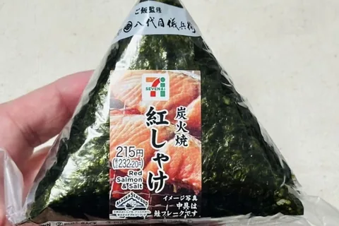 A 7-Eleven charcoal-grilled red salmon onigiri held in a hand; the label reads 215 yen
