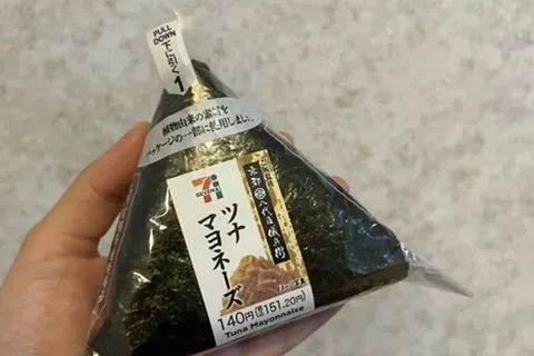 A 7-Eleven hand-rolled tuna mayonnaise onigiri held in a hand; the label reads 140 yen