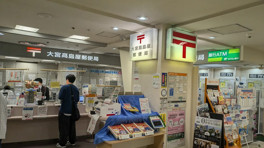 埼玉县大宫高岛屋邮局的柜台,上方挂着红色邮局标志“〒”和邮局名称招牌,旁边有ATM的照片