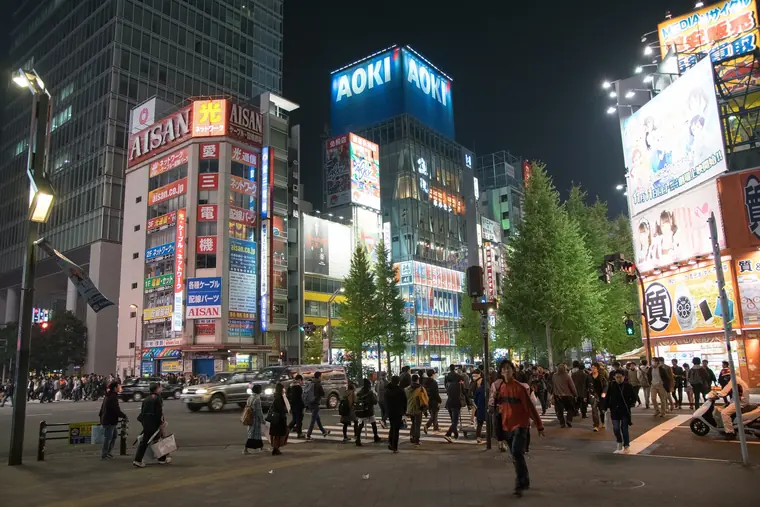 Pedestrians crossing Chuo-dori in Akihabara at night under towering electronics-store signs and anime billboards.