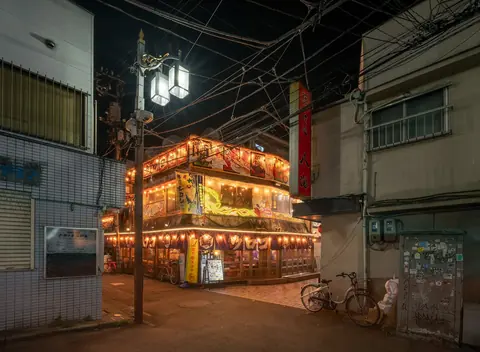 A two-story izakaya in Nakano, Tokyo, glowing with lanterns and string lights on a quiet street at night.