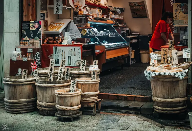 A food shop at a Kyoto market with wooden barrels of pickles and handwritten price tags.