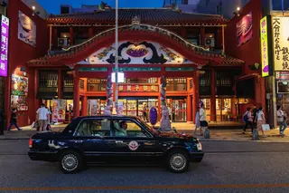 A black taxi passing a red Okinawan-style shopfront on Kokusai-dori in Naha at night.