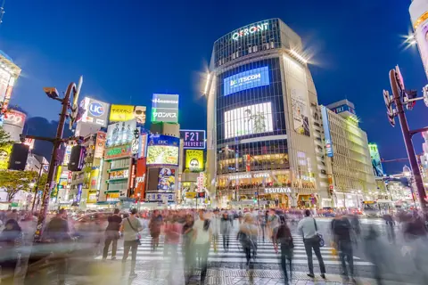Pedestrians blurred in motion on the Shibuya Scramble Crossing under giant video screens at blue hour.