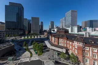 The red-brick Marunouchi building of Tokyo Station and its central plaza, seen from above, surrounded by glass office towers under a clear blue sky.