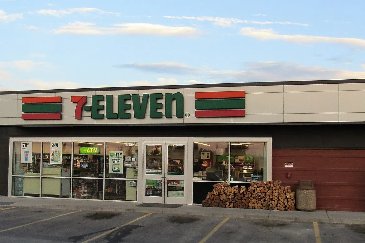 Exterior of a 7-Eleven convenience store in the United States with red and green striped signage, a parking lot, and stacked