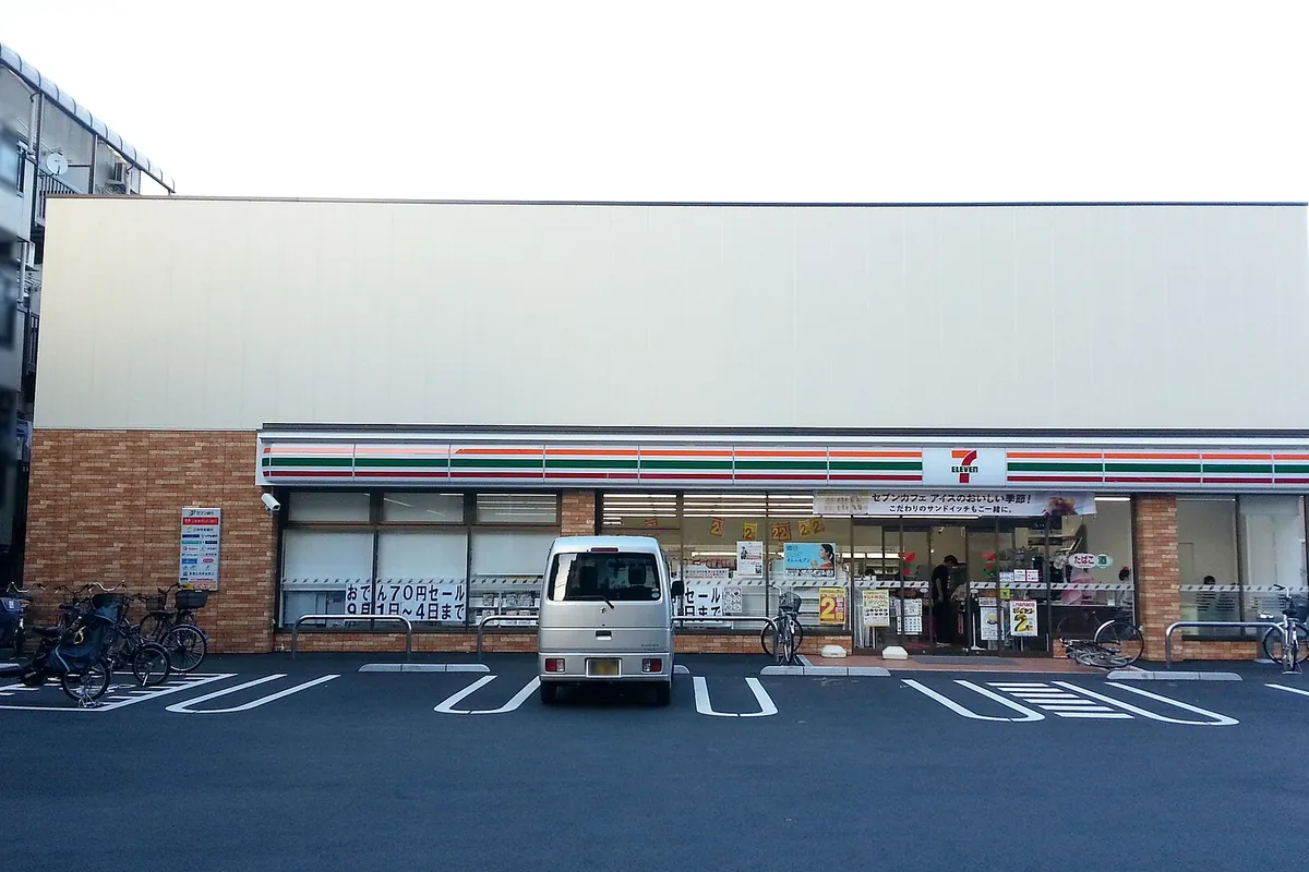 Exterior of a 7-Eleven convenience store in Tokyo with bicycles parked on both sides and a small van parked in front
