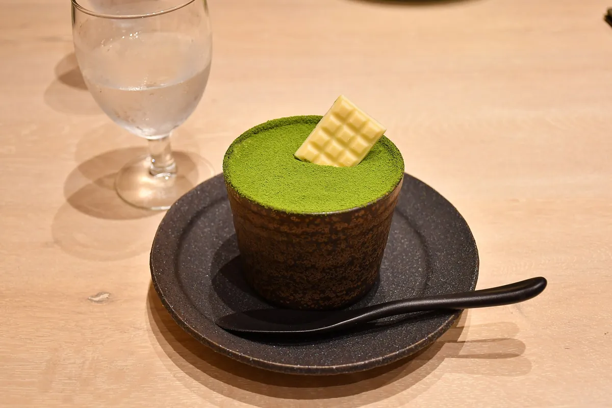 A matcha tiramisu dessert on a dark plate with a spoon, next to a glass of water on a wooden table