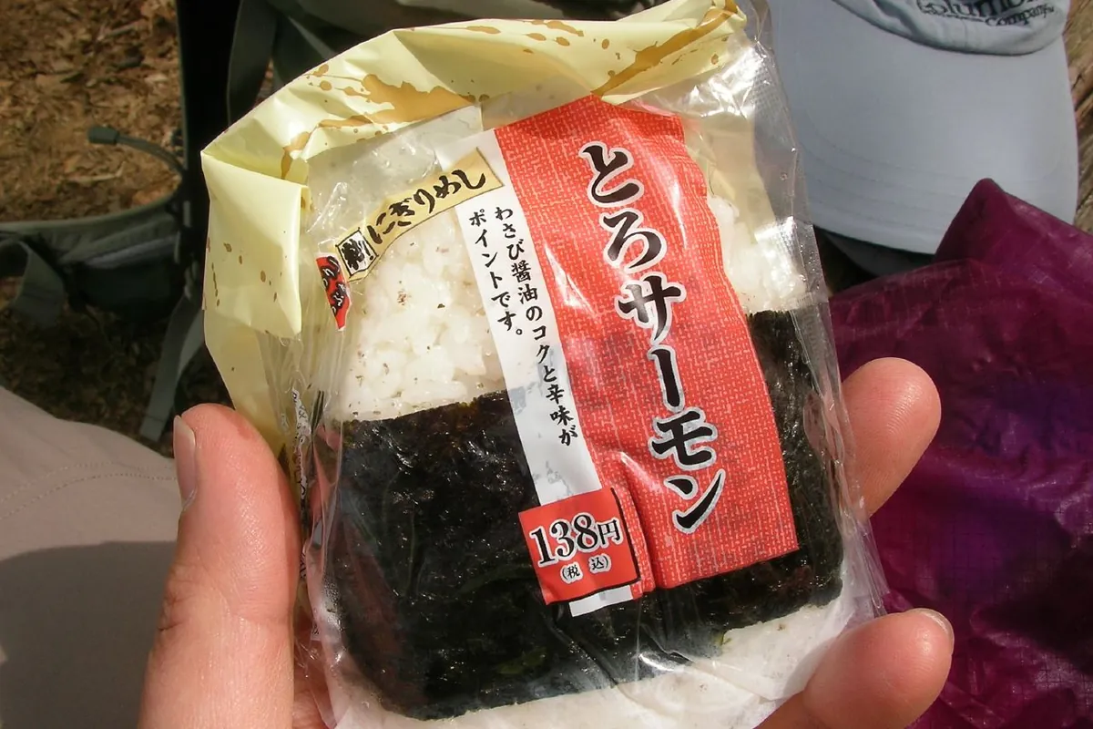 手上拿著一顆用海苔包著的包裝鮭魚飯糰，貼有紅白標籤與價格標籤