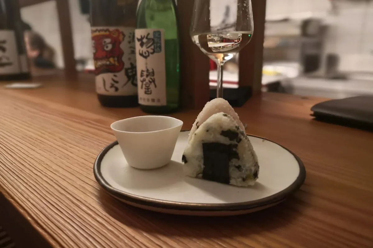 盤子上的三角飯糰，旁邊有一個小杯子和一杯清酒，木頭吧檯上擺著清酒瓶