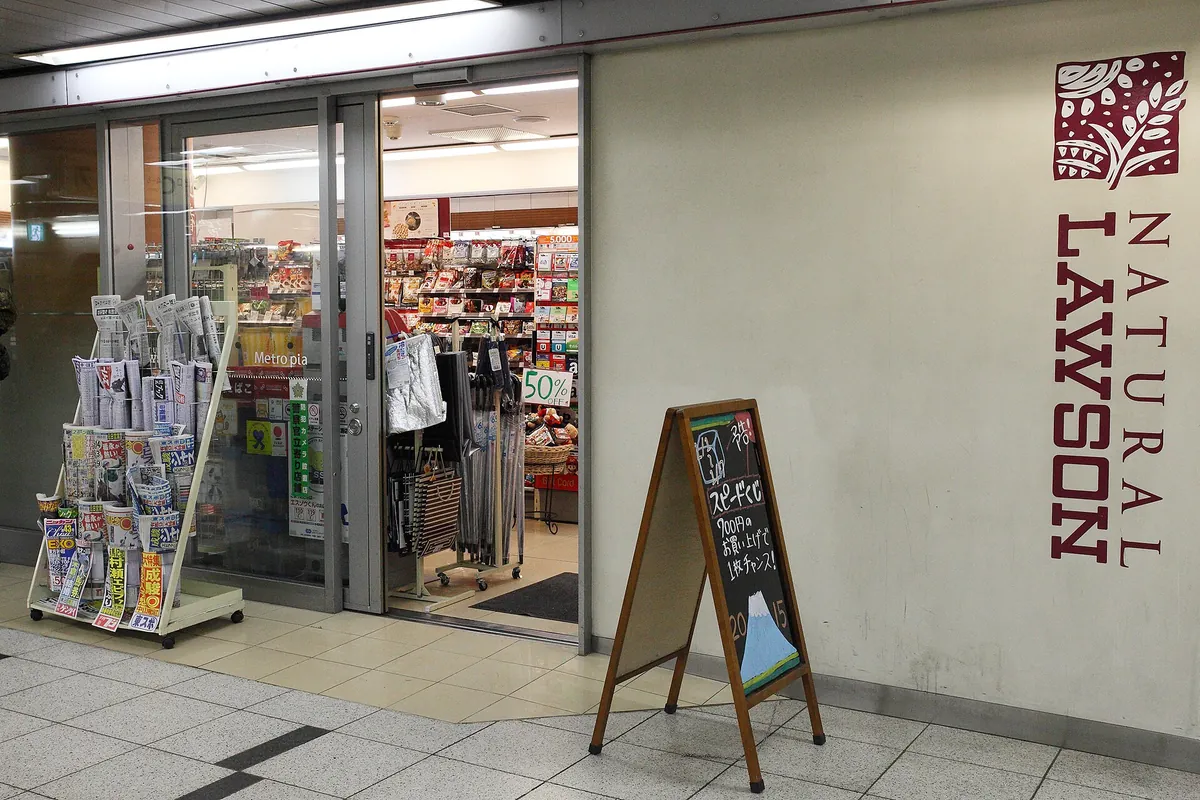 Entrance of a Natural Lawson convenience store with a newspaper rack, sandwich board sign, and open sliding doors