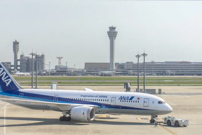 An ANA Boeing 787 parked at the gate with a tow tug in front, an airport control tower and terminal buildings behind