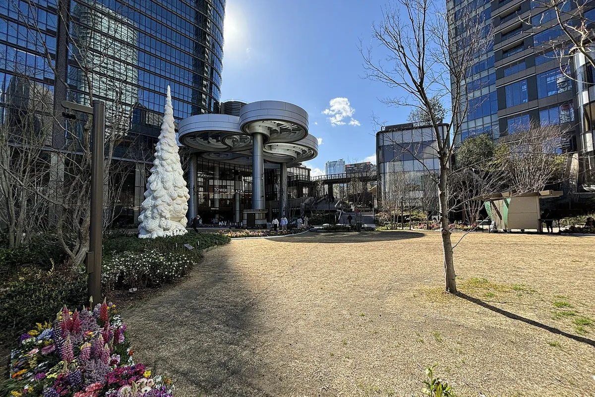 麻布台之丘的草地廣場，有白色圓錐形的樹木雕塑、落葉的樹、花圃，後方是高樓
