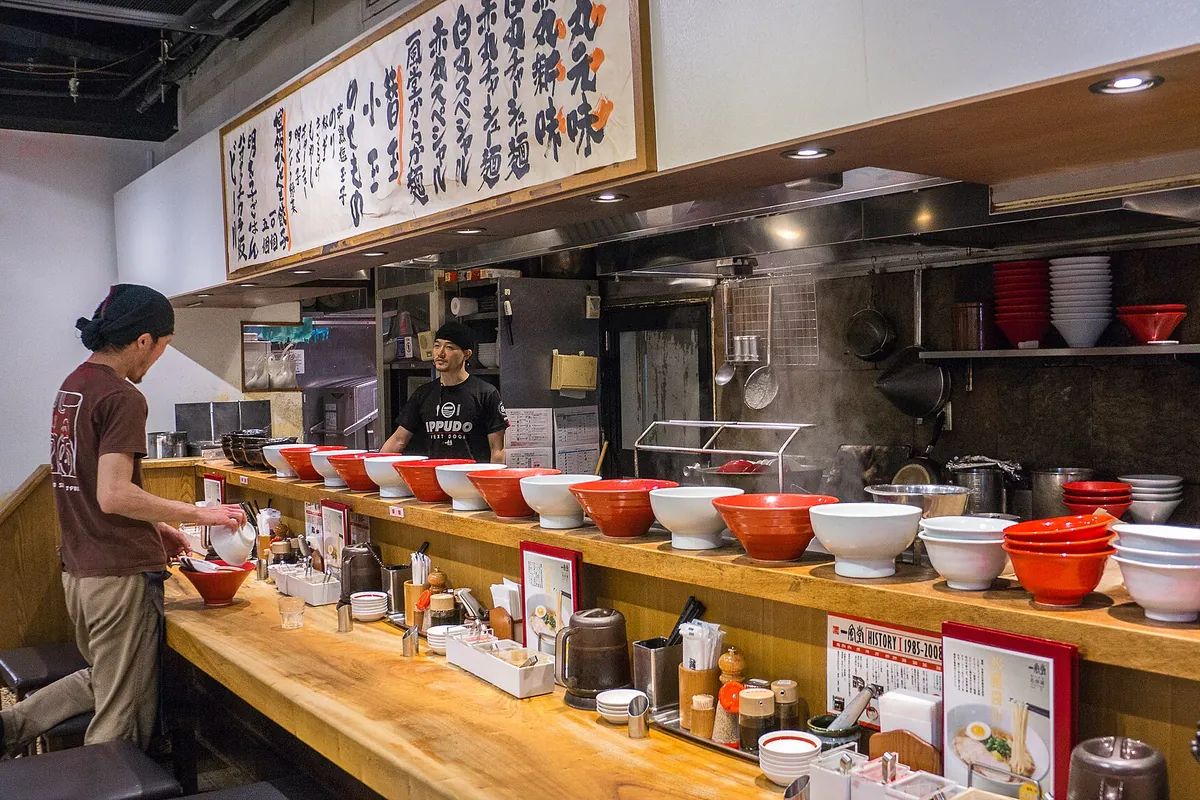 A ramen shop counter at Ippudo in Tokyo lined with red and white bowls, with staff preparing noodles behind it