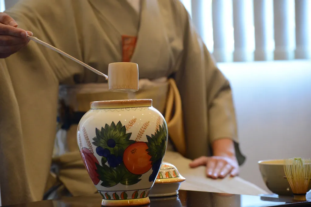 A hand in a kimono sleeve holding a bamboo tea scoop above a fruit-patterned ceramic tea caddy, with a tea bowl nearby