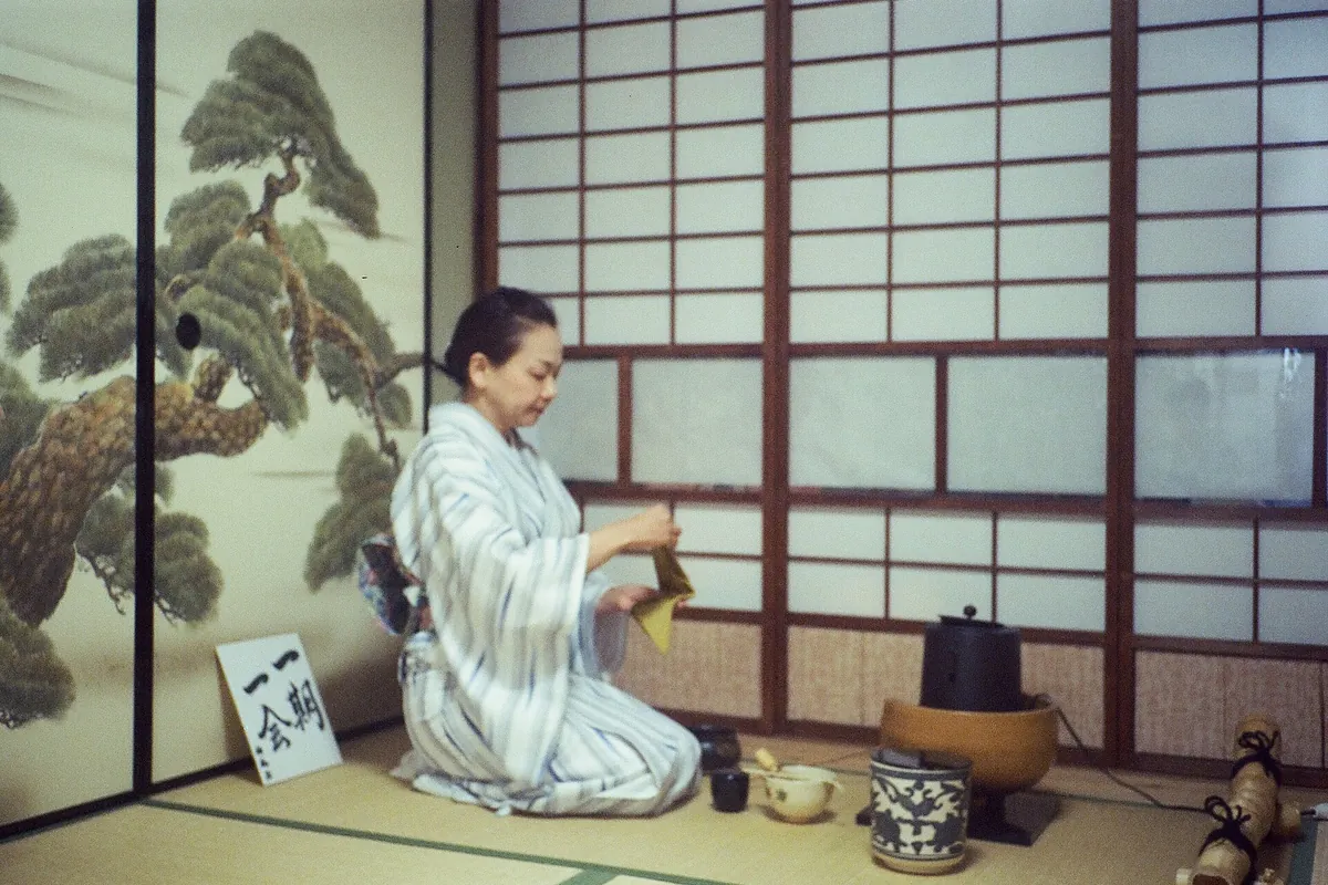 A kimono-clad woman kneeling on tatami folding a yellow cloth with an iron kettle on a brazier and pine-painted screen behind