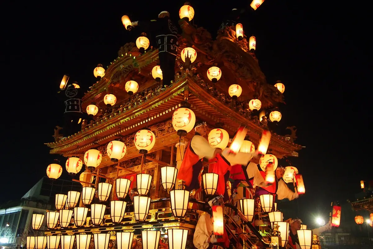 Ornate festival float with a gold-trimmed carved roof, rows of round paper lanterns, and people in festival robes riding on i