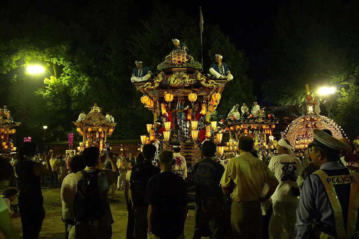 Crowd watching illuminated festival floats at night, with a police officer in a POLICE vest standing in the foreground