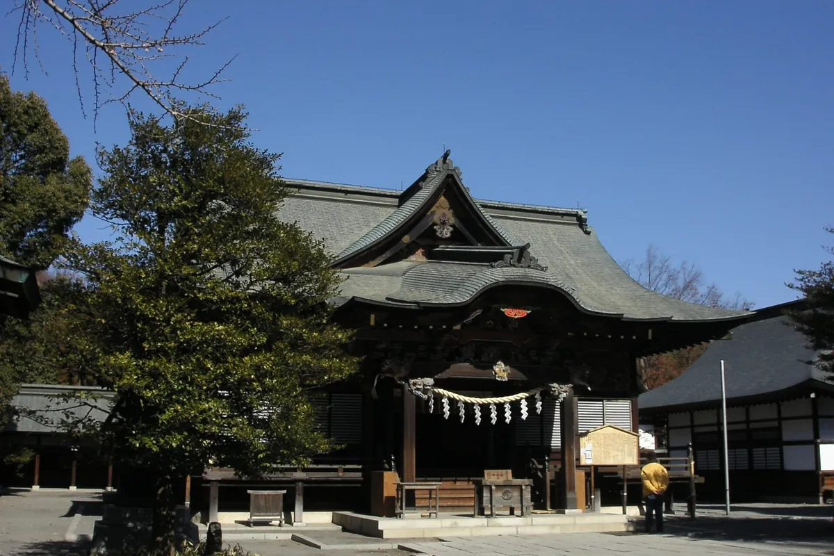 Main hall of a wooden Shinto shrine with a dark tiled roof and a shimenawa rope hanging at the entrance, surrounded by trees