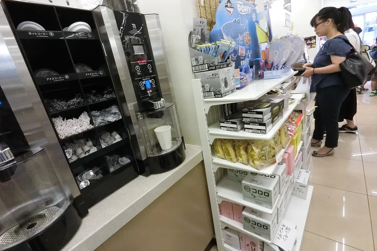 コンビニのレジ横にあるセルフサービス式のコーヒーマシンとカップの受け取り口、近くに商品棚がある様子