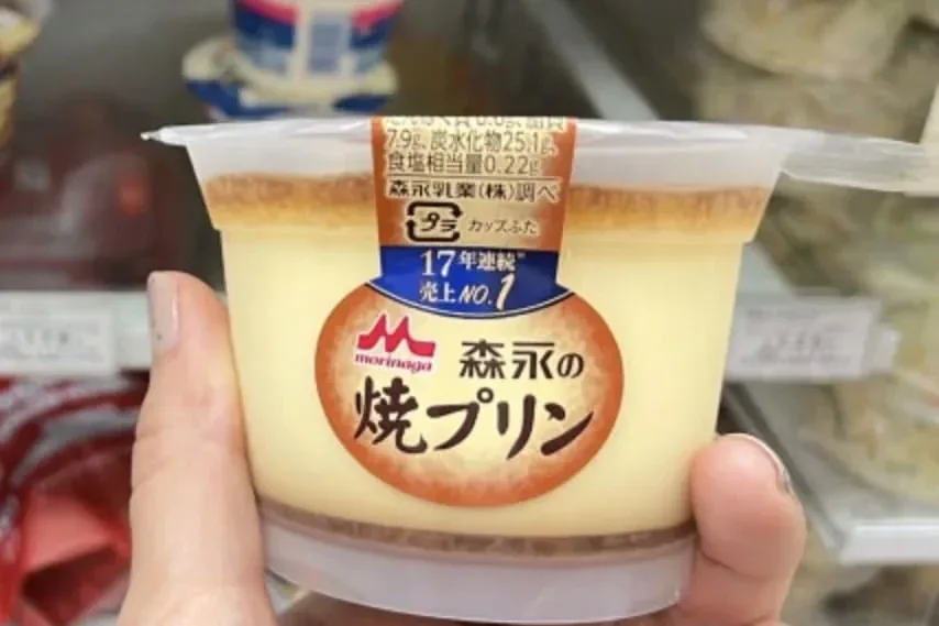 A Morinaga baked custard pudding cup held up in front of a convenience store chiller shelf in Tokyo