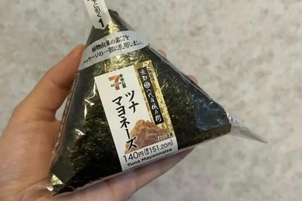 A 7-Eleven tuna mayonnaise onigiri in its wrapper, with a label of 140 yen (151.20 yen with tax), held in a hand