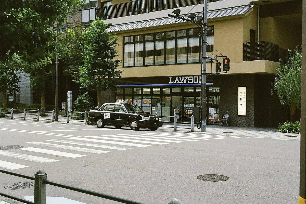 The storefront of a Lawson convenience store in Tokyo, with a black taxi parked outside and a crosswalk in front.