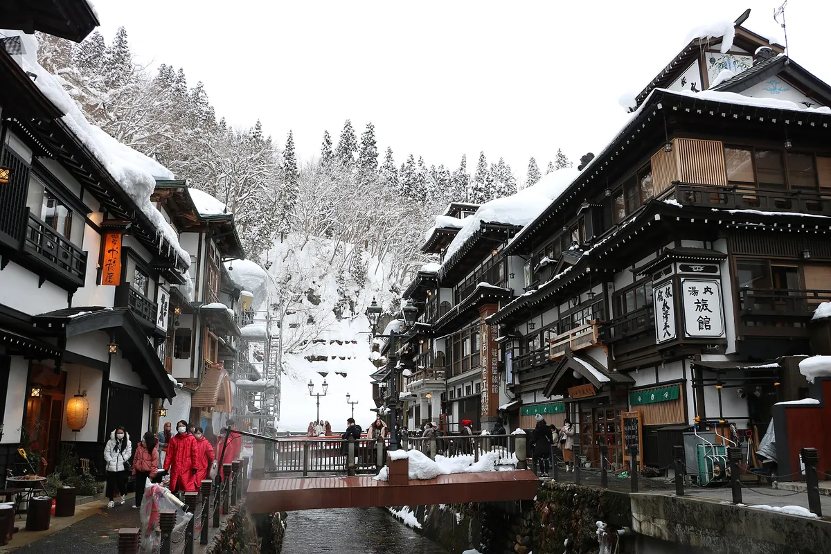 銀山温泉で川沿いに並ぶ雪に覆われた木造の多層旅館。冬のコートを着た人たちが橋を渡っている