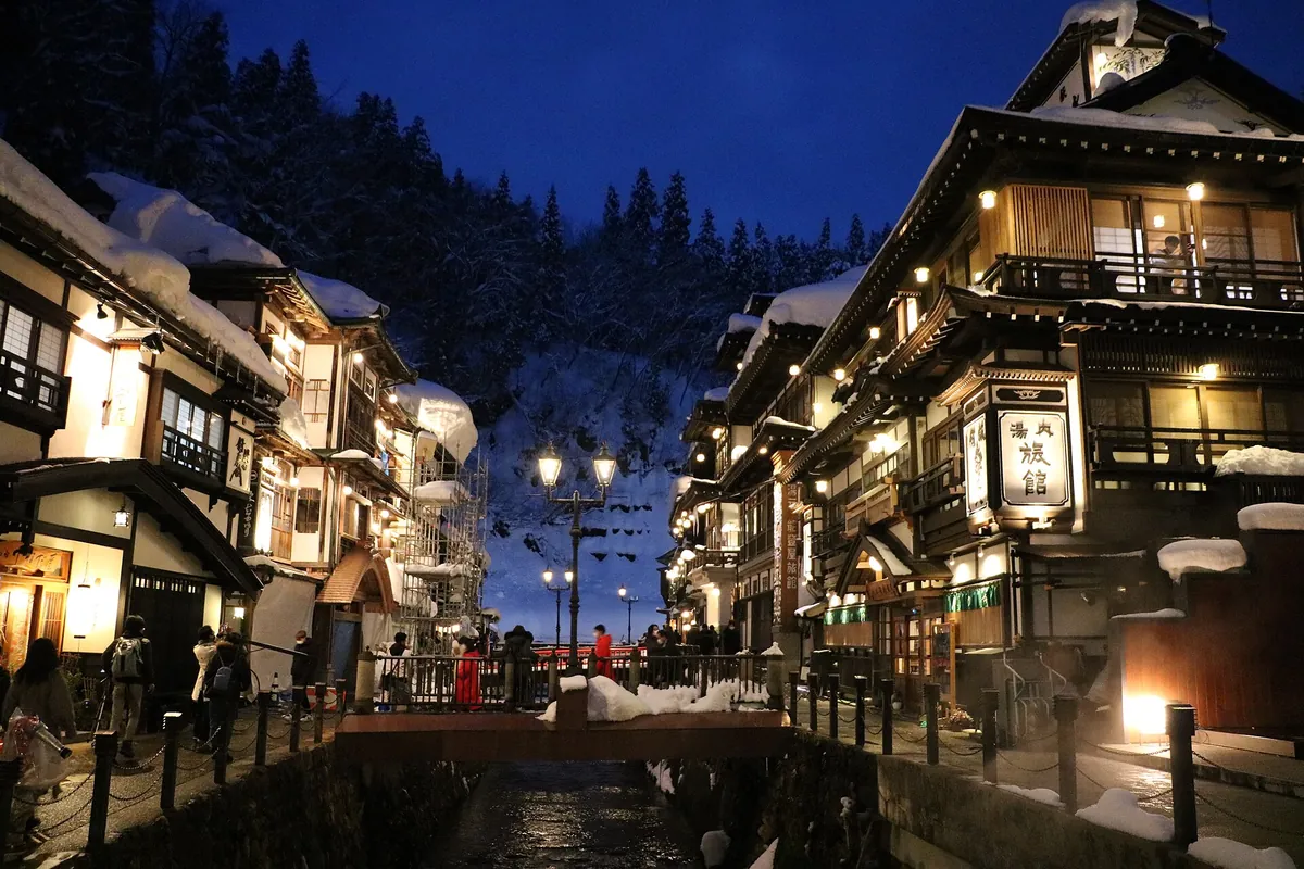 夕暮れ時に川沿いで明かりがついた銀山温泉の旅館。橋の上にガス灯があり、来訪者が欄干の近くに集まっている