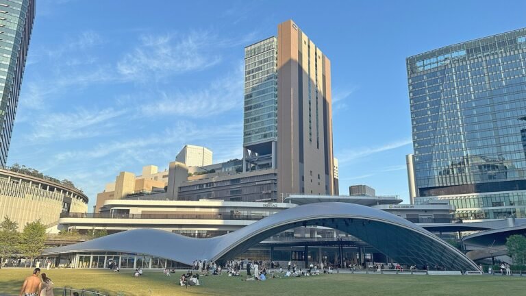 Grand Green Osaka's curved glass pavilion and lawn with skyscrapers behind, people relaxing on the grass