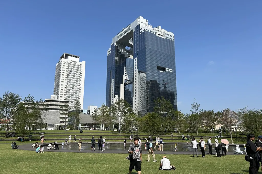 Twin glass towers connected by a circular rooftop deck at Grand Green Osaka, with a lawn, fountain and people walking below