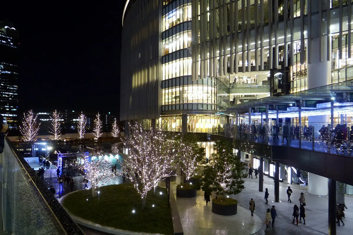 Trees strung with white string lights outside Grand Front Osaka at night, with the building's terrace and people walking