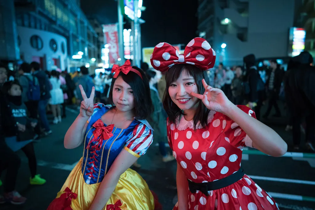 Two women dressed as Snow White and Minnie Mouse making peace signs on a crowded Shibuya street at night