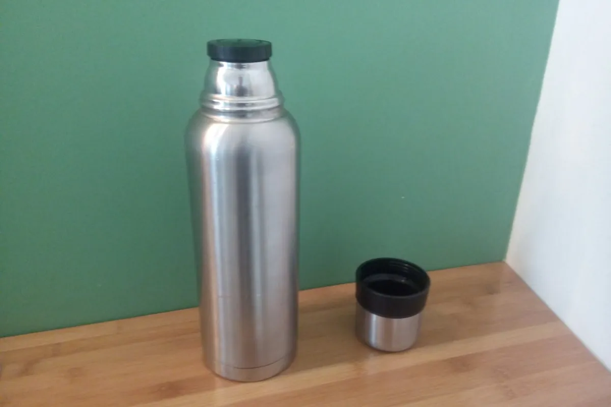 A stainless steel vacuum flask with a black cap, and its cup lid resting beside it on a wooden surface
