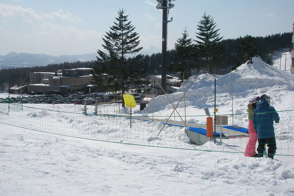 Two people in ski gear near a snowy slope with a moving walkway, parked cars, and resort buildings