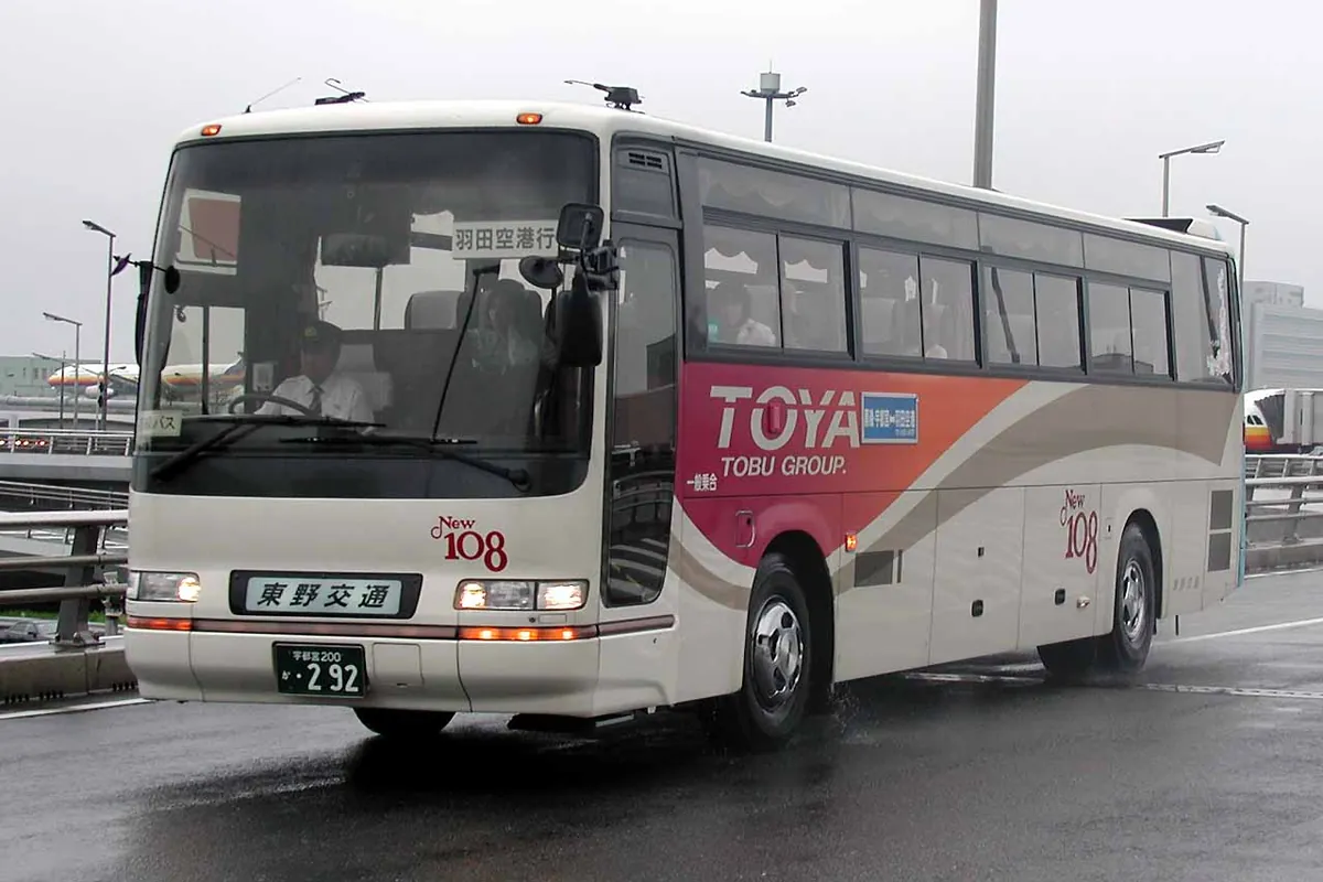 A white bus with an orange and red TOYA livery and a Haneda Airport destination sign driving on a wet road near the airport