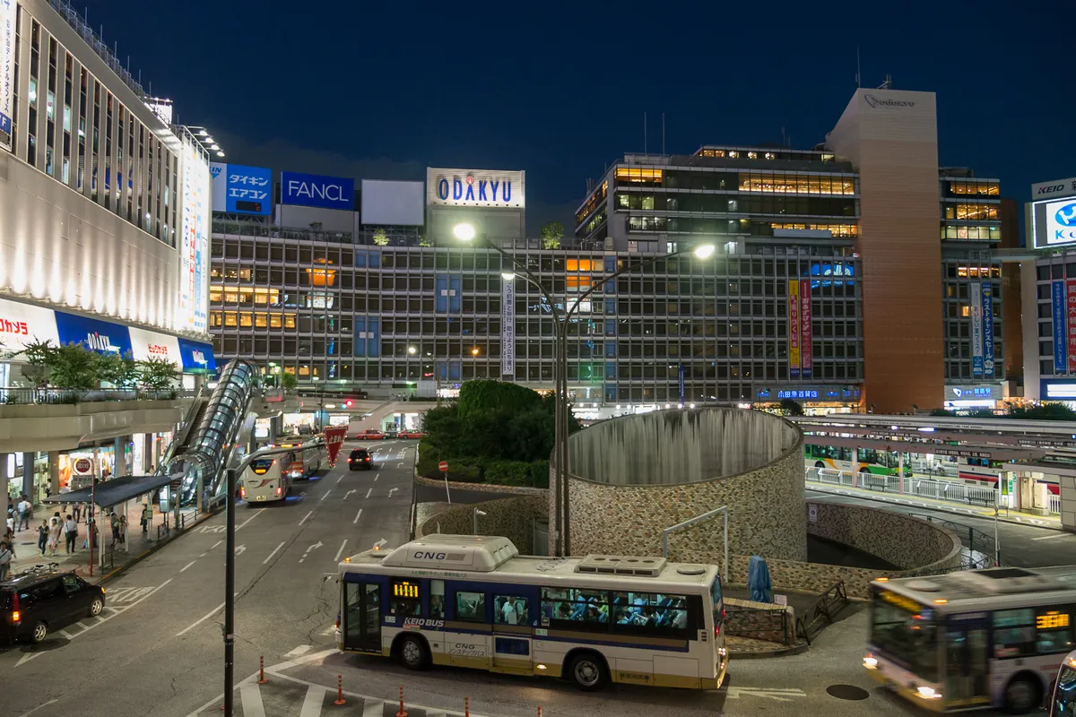 Shinjuku Station's west exit plaza at night, with illuminated department store buildings and buses at the bus terminal