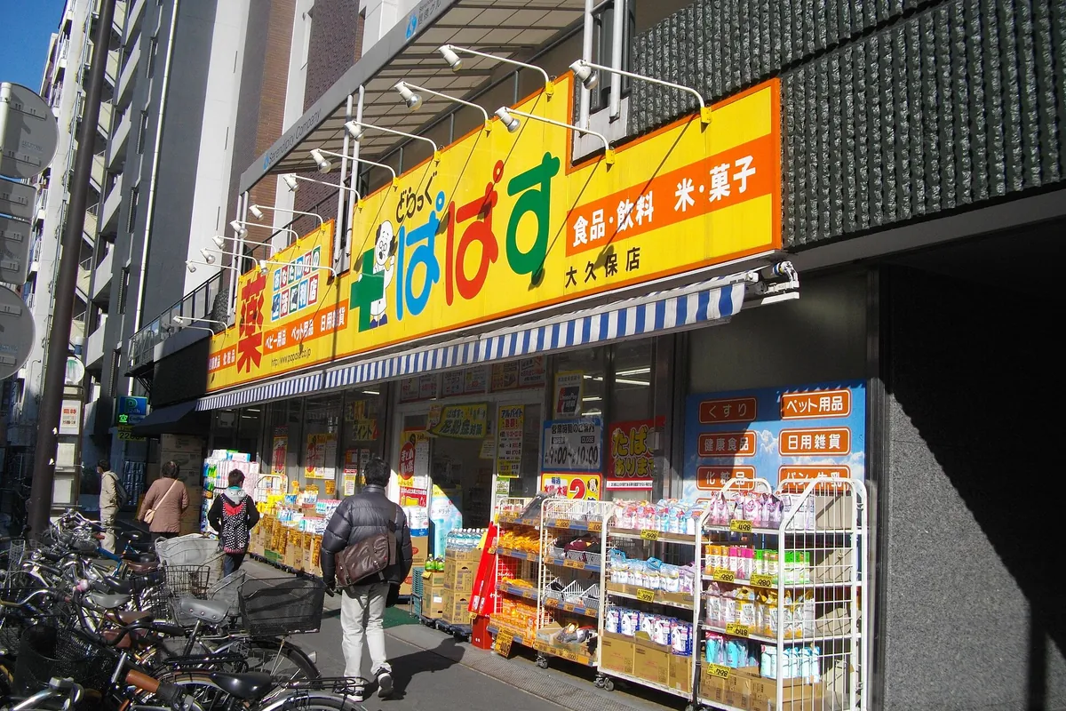 Exterior of a Japanese drugstore with a yellow sign in Japanese characters, bicycles parked outside, and goods displayed on t