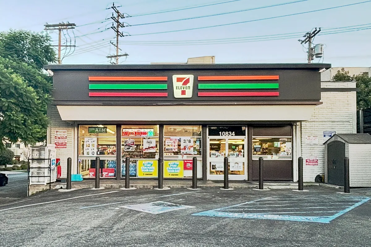 A 7-Eleven convenience store with red, orange, and green striped signage and a parking lot with bollards in West Los Angeles