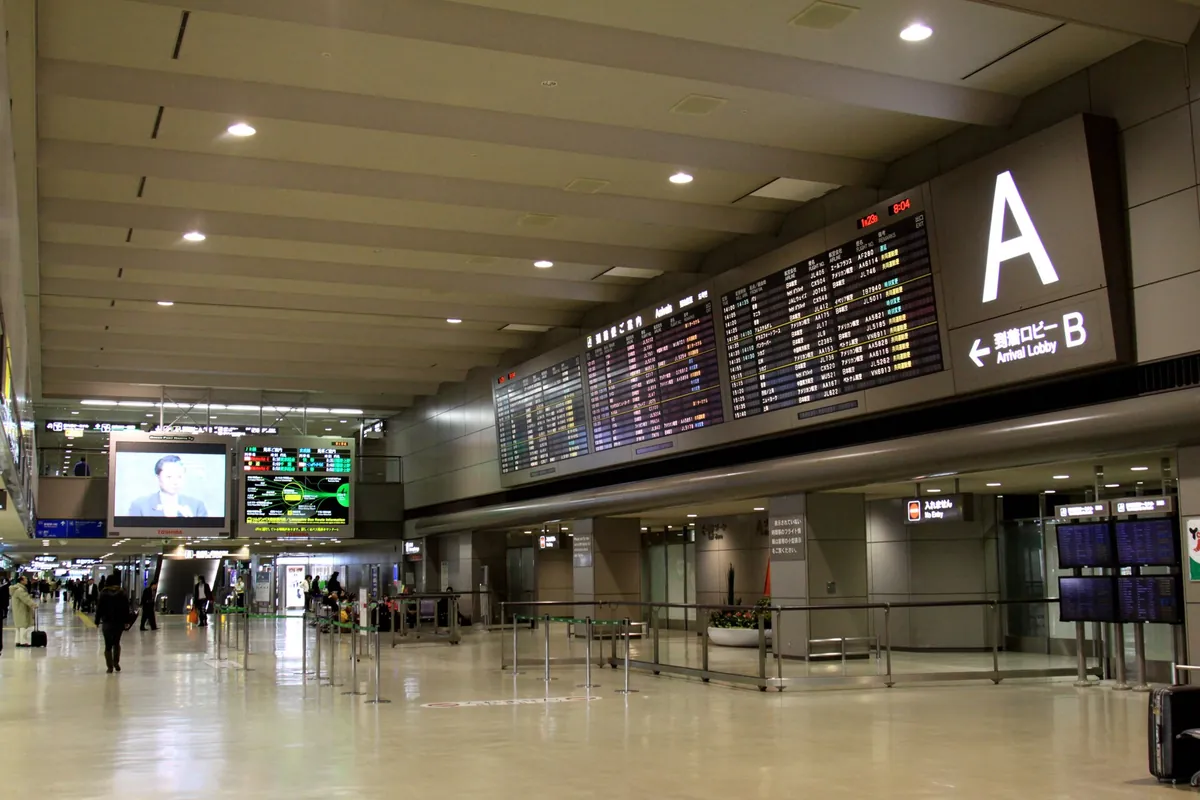 Airport terminal hallway with a large flight information board and Arrival Lobby signage in Japanese and English, people walk