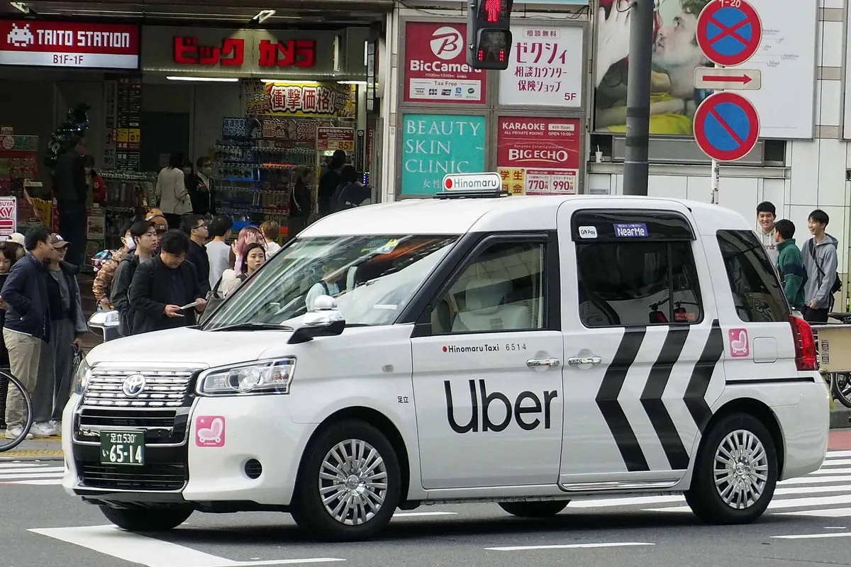 Uberのロゴと黒いシェブロン柄が入った白いトヨタJPN TAXIが、東京のビックカメラの前で人混みのそばに停まっている