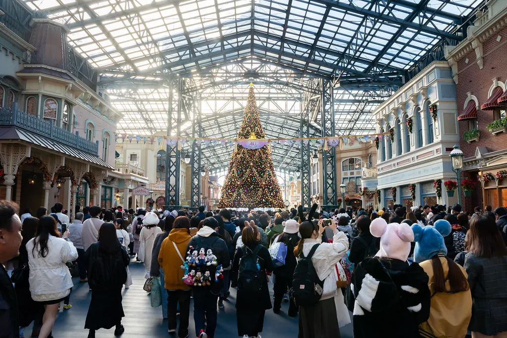Crowds walking under the glass-roofed World Bazaar arcade at Tokyo Disneyland, with a large Christmas tree ahead