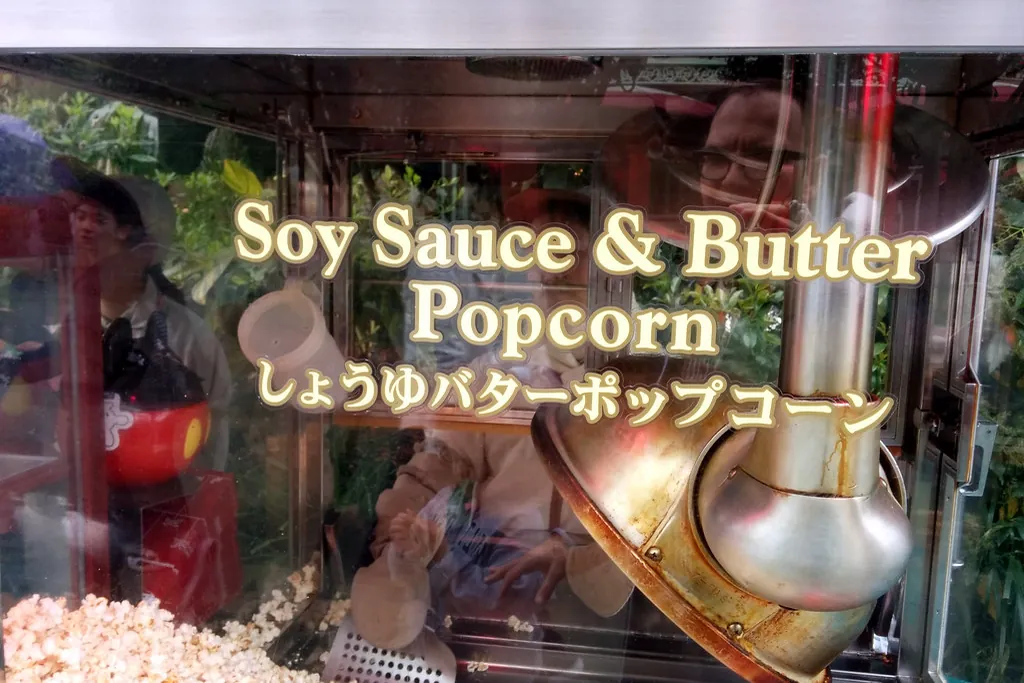 A popcorn wagon window with soy sauce and butter popcorn signage in English and Japanese, popcorn visible below