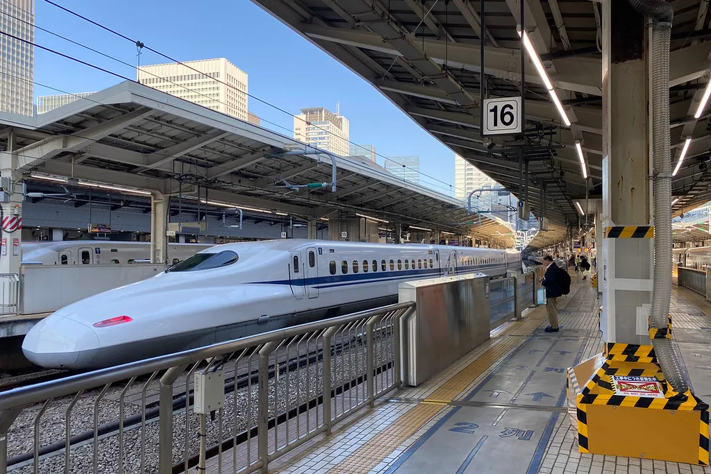 A white N700 series Shinkansen train stopped at platform 16 under a station canopy with city buildings behind it