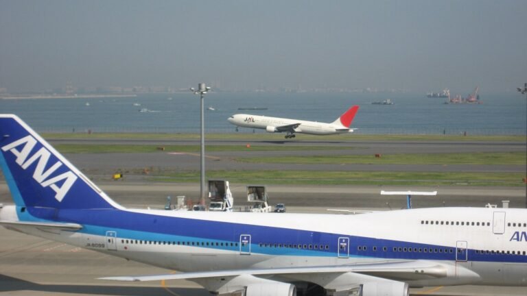An ANA Boeing 747 tail and fuselage with a JAL aircraft taking off in the background at Haneda Airport