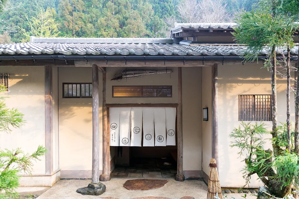 Wooden entrance of a traditional ryokan with white noren curtains, surrounded by trees and hills